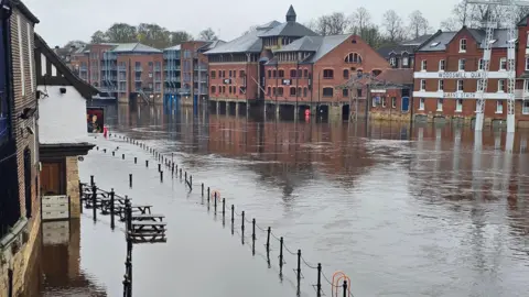 BBC Weather Watchers/Gryning Flooded riverside area in York with water covering the walkway and reaching the ground floors of buildings. Several brick buildings, including converted warehouses and apartments, stand along the riverbank. Metal railings and benches are partially submerged.