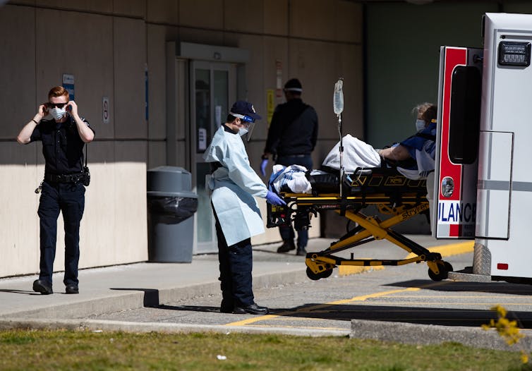 An EMT wearing a face mask rolling a gurney out of an ambulance while another EMT puts on a face mask in the background.