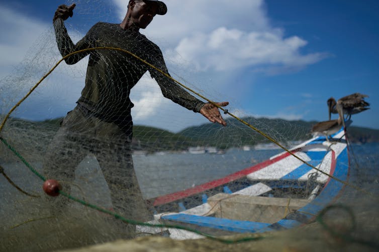 A fisherman carries a net, he is standing next to a small boat near a body of water