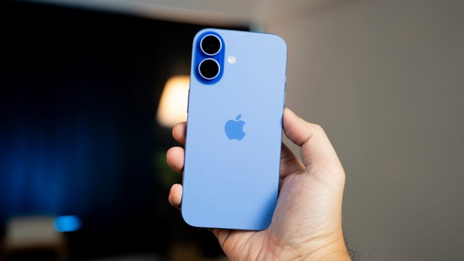 Hand holding a blue iPhone with dual rear cameras and Apple logo visible, against a blurred indoor background.
