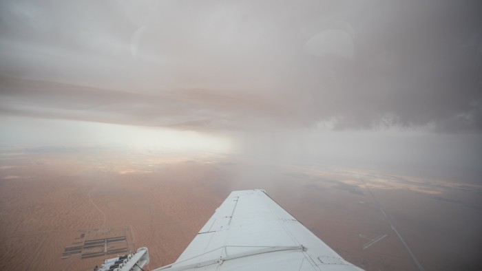 A view from a plane wing shows rain falling from dark clouds over a desert landscape during a cloud-seeding mission.