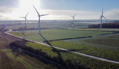 Several large wind turbines stand in open fields at an onshore wind farm under a clear sky.