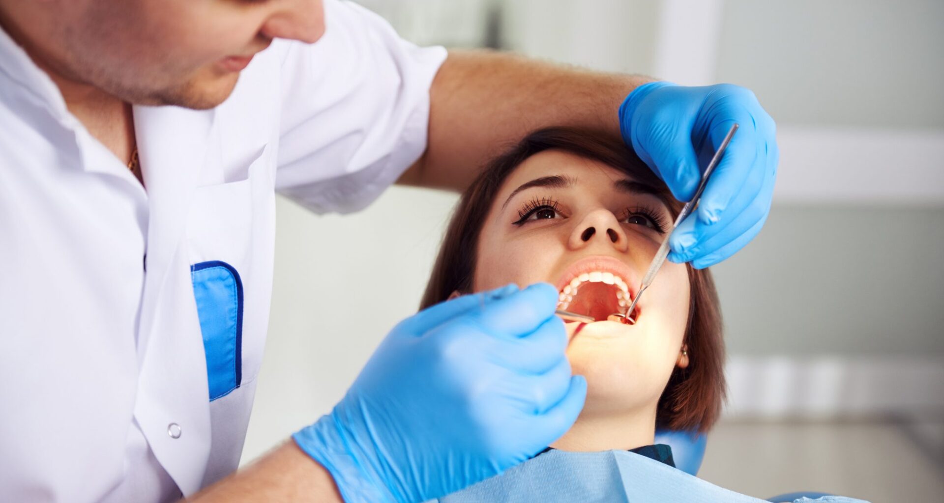 Male doctor in uniform checking up female patient's teeth in dental clinic. Concept of oral examination, toothache and decay treatment.