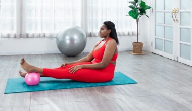 woman wearing red leggings and crop top sitting with legs outstretched on a turquoise exercise mat. there's a pink pilates ball under one foot and she's in a studio setting.