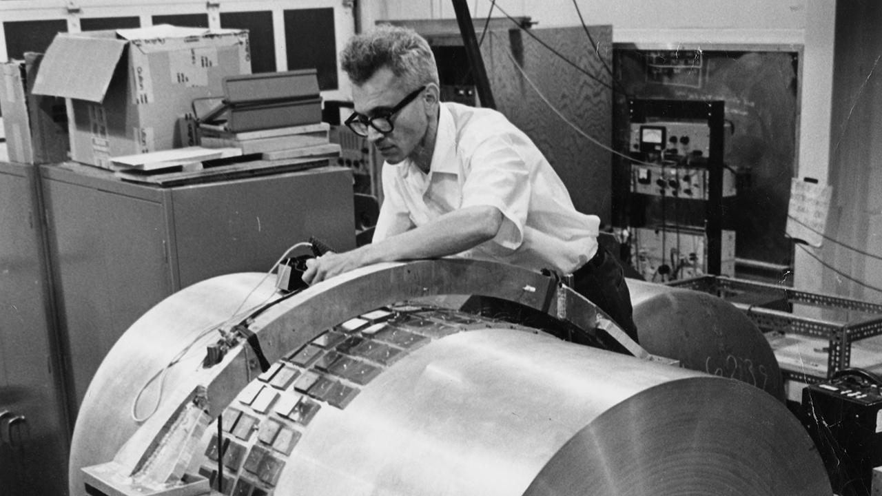 A man adjusts components on a large, cylindrical piece of scientific equipment—resembling early Weber bars used to detect gravitational waves—in a laboratory setting.