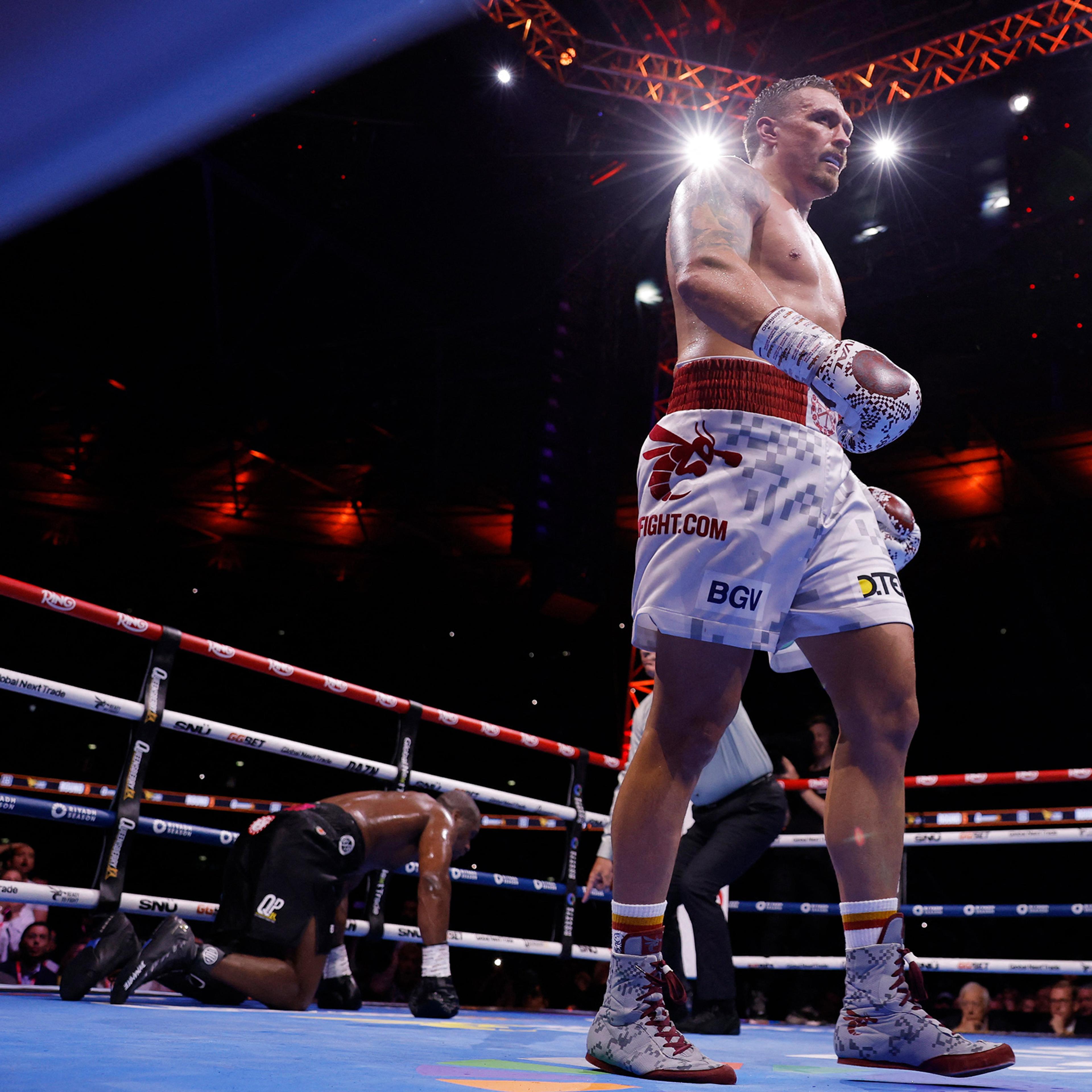 Photo of a boxing match with a standing boxer in the ring and an opponent kneeling behind, lit by overhead lights.