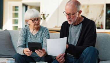 A senior couple sitting on a couch in front of a table with papers on it. The man is holding up a piece of paper that they are both looking at