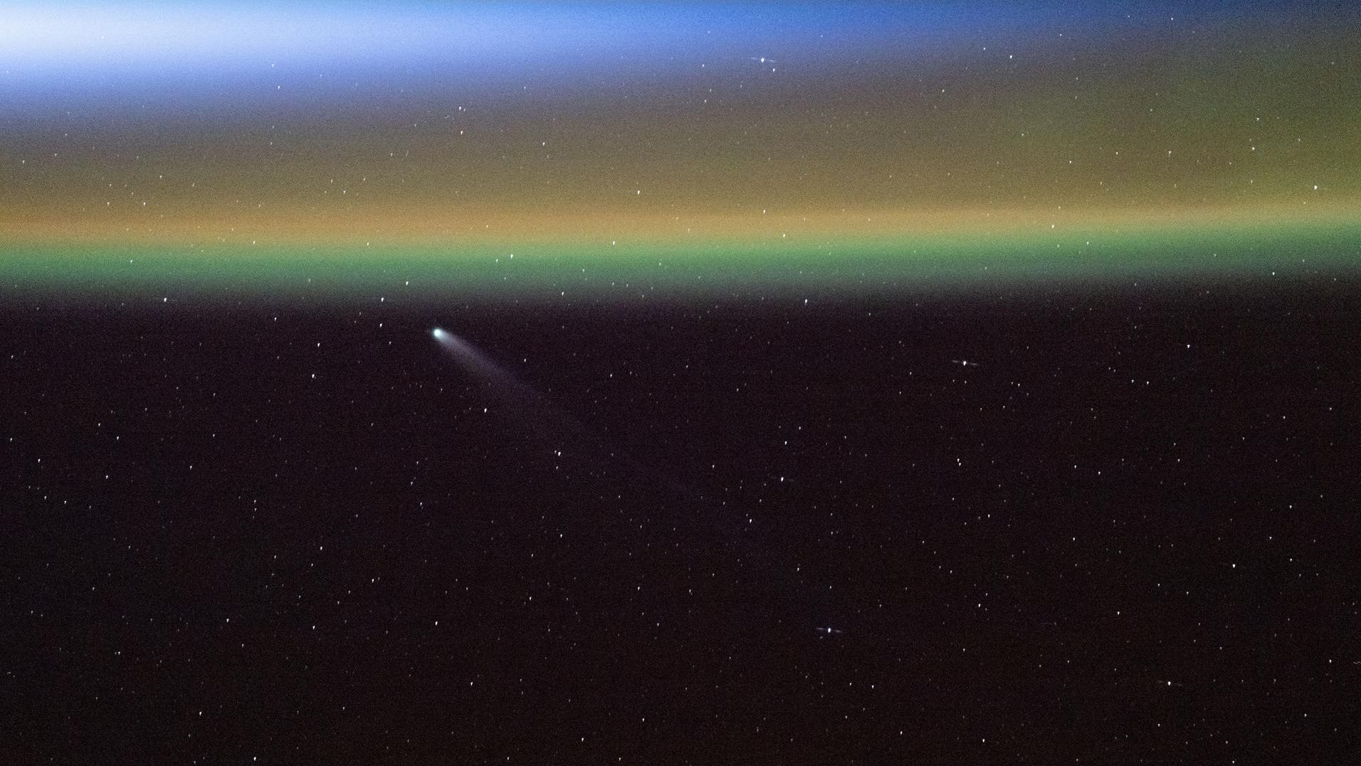 A streak of white against the darkness of space shows comet Swan moving toward the top left of the image near a series of yellow, green and blue stripes of the northern lights appearing on Earth
