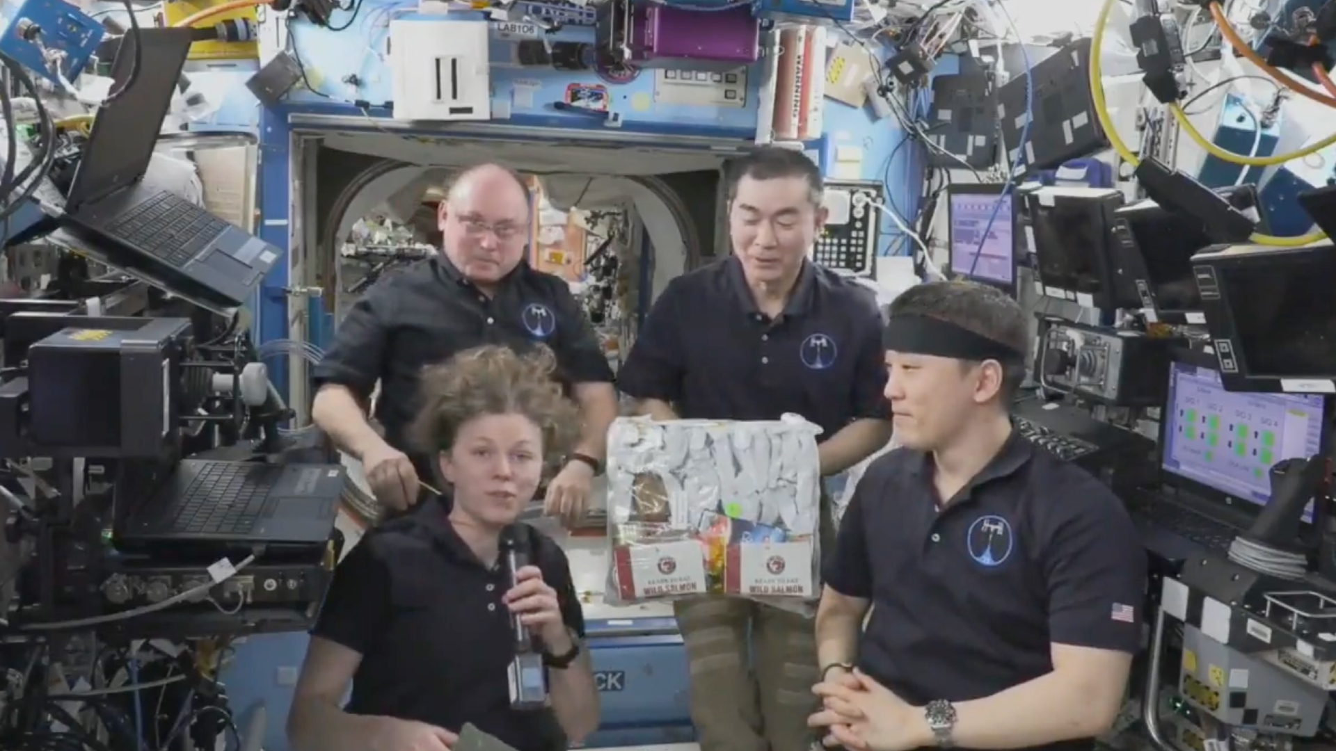 Four astronauts wearing black shirts float around a bag of prepackaged food in the International Space Station