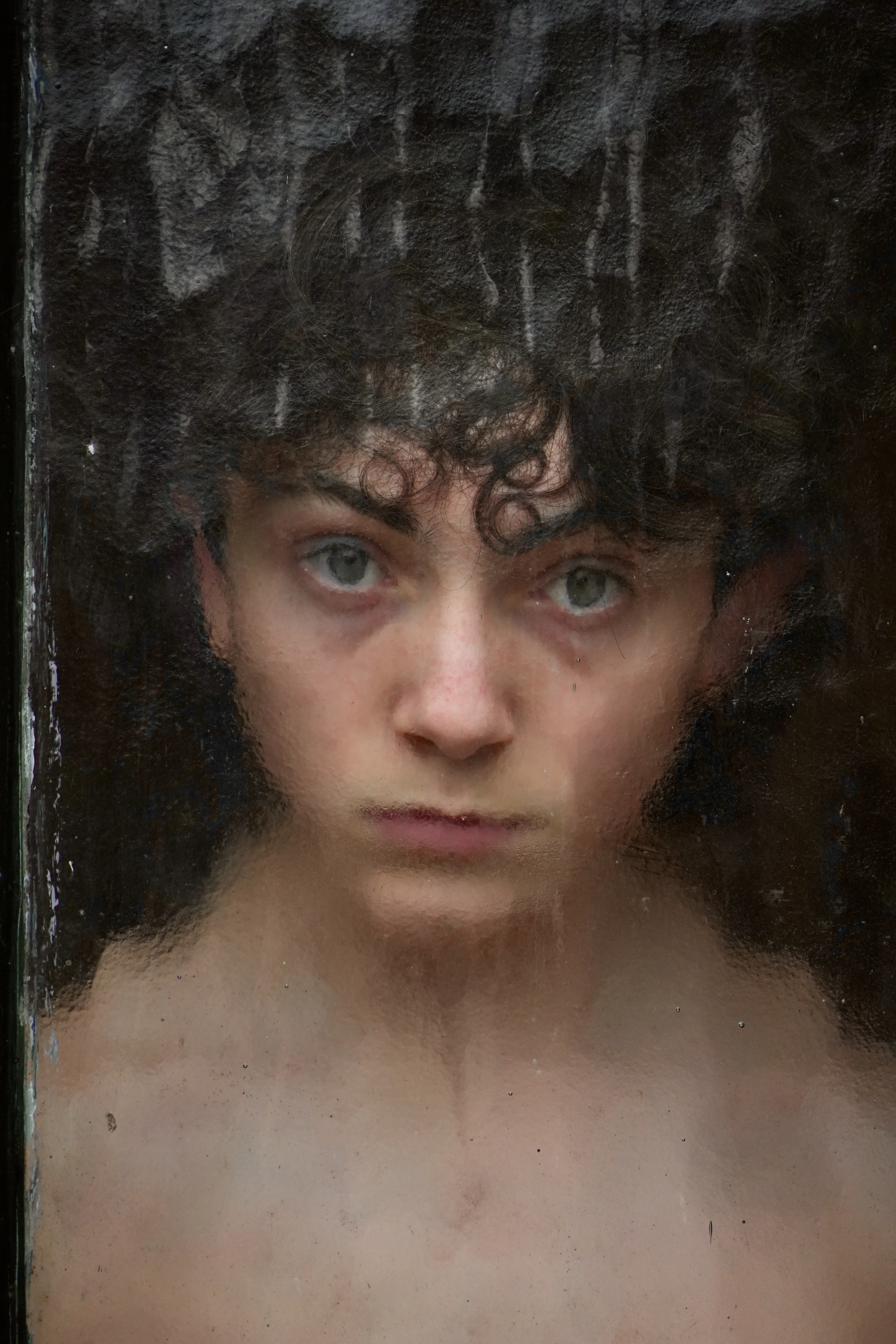 Portrait of Peter, a person with dark curly hair and light eyes, behind textured glass.