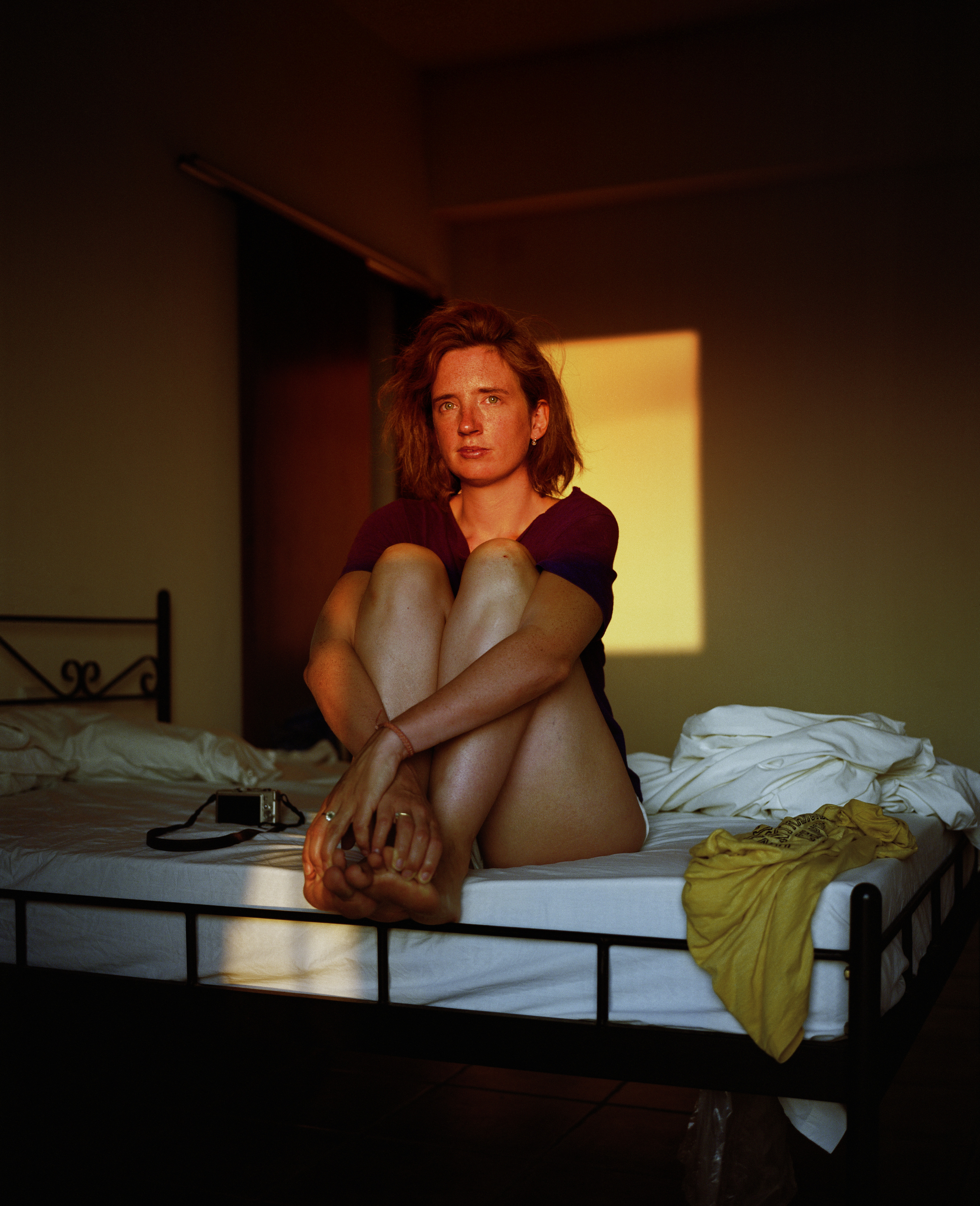 A woman with red hair and freckles sitting on a bed with knees hugged, looking at the camera.