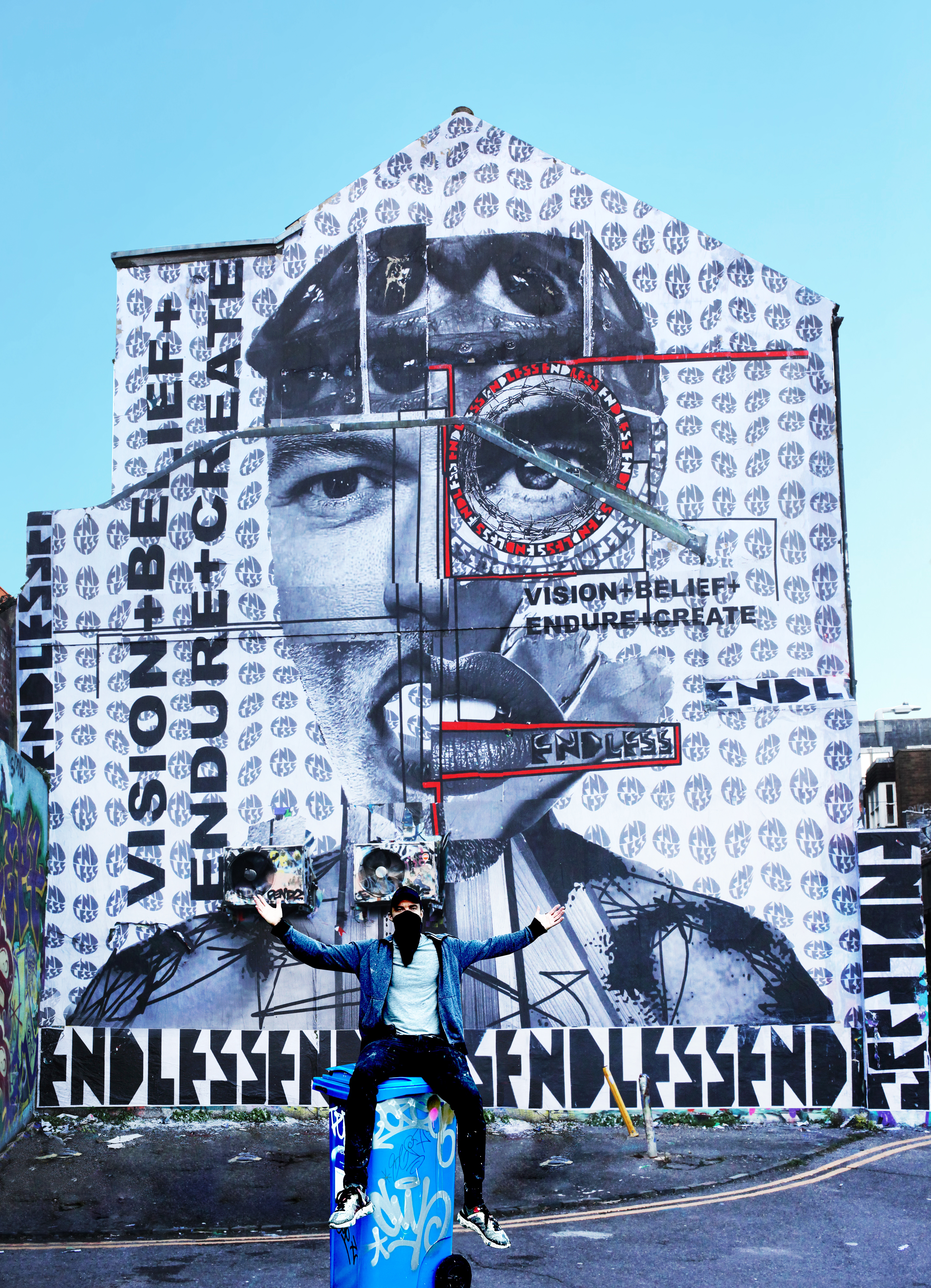 Artist Endless sitting on a blue bin in front of a large-scale mural in Brighton featuring a portrait of Luke Evans and the words "Vision + Belief + Endure + Create."