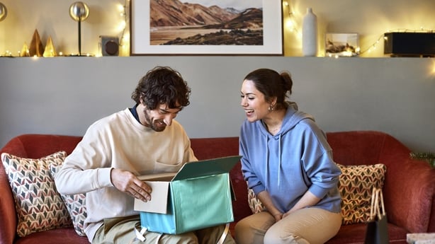 Surprised boyfriend opening gift given by girlfriend. Happy couple is celebrating Christmas at home. They are sitting on sofa.