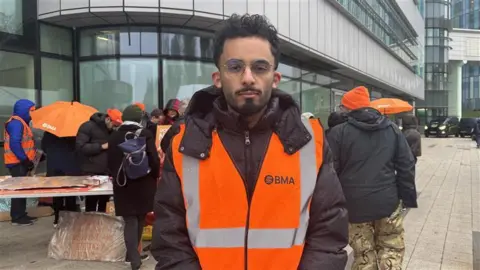 A man with black curly hair and facial hair wearing circular glasses. He is standing outside a glass hospital and is wearing a black puffer coat and an orange hi-vis jacket