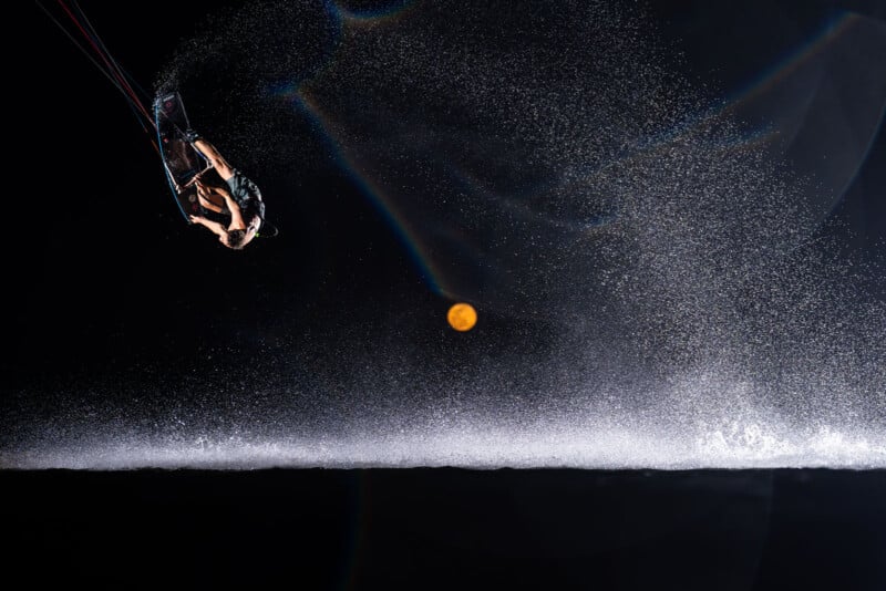 A wakeboarder performs an aerial trick at night, illuminated by a bright flash. Water sprays dramatically in an arc, and a full moon glows in the dark sky behind, creating a dynamic and striking scene.
