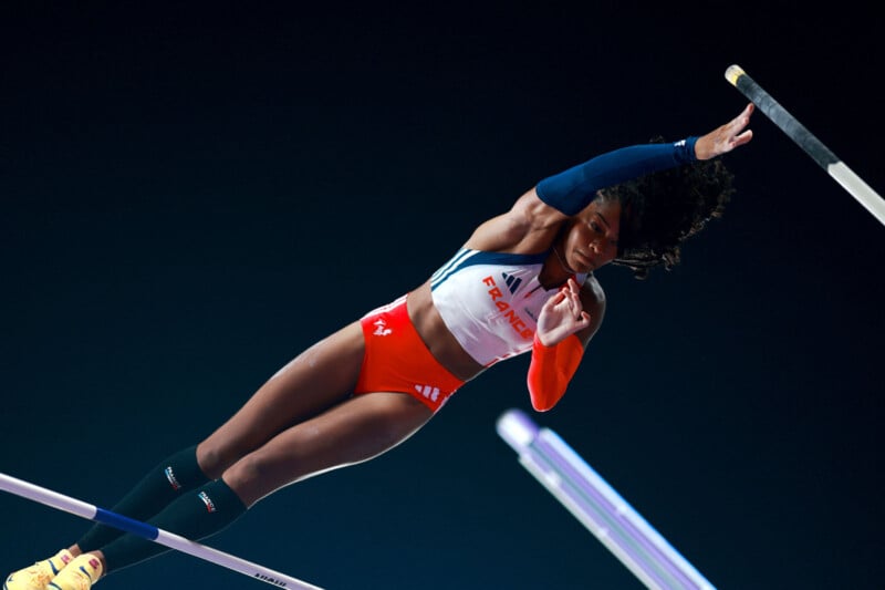 A female pole vaulter in mid-air, wearing a red and white uniform and black knee-high socks, clears a pole against a dark background during a competition.