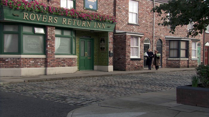 Ken and Rita, linking arms, carry flowers along Coronation Street beside the Rovers Return