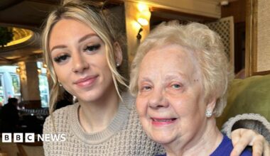 A picture of a female in her 20s with her arm around her grandmother. The younger female has blonde hair which it is tied up in a ponytail wearing a tan knitted jumper. She is wearing diamond studded earrings. The elderly lady on the right has blonde permed hair she is wearing a blue knitted jumper also she has a pair of diamond studded earrings.