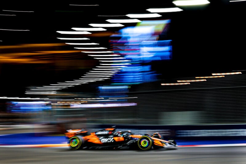 A Formula 1 car speeds along a brightly lit night track, with motion blur emphasizing its fast movement and trailing lights in the background.