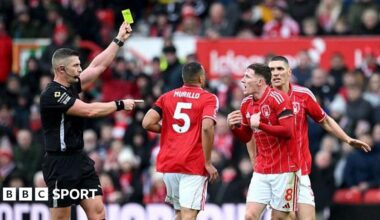 Referee Rob Jones books Elliot Anderson, with the official raising his yellow card above head height and pointing towards the animated Nottingham Forest player, in red and white, who has both hands out in front of his chest