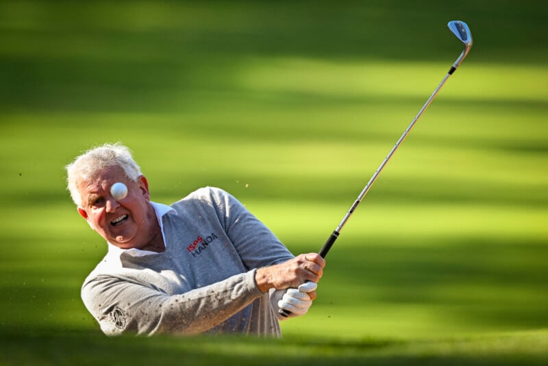An older man playing golf swings his club, sending the golf ball into the air out of a sand bunker on a bright, sunny day, with a green field in the background.