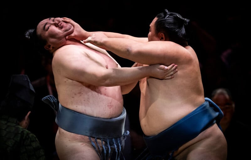 Two sumo wrestlers compete in a match, one pushing the other's face with an outstretched hand. Both wear traditional mawashi belts, and the background is dark, focusing attention on the wrestlers’ intense struggle.