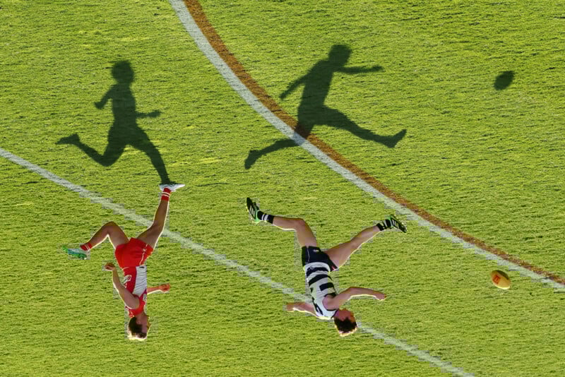 Two Australian rules football players, one in red and one in black and white stripes, run and kick on a grassy field, casting long shadows in the sunlight. A yellow ball is visible near the player in stripes.
