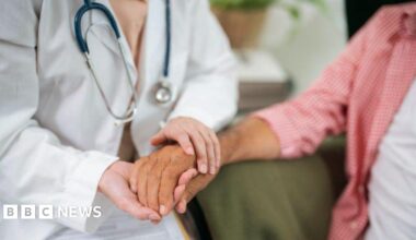 A doctor holds a woman's hand in a consultation. The doctor has a white coat and a stethoscope.