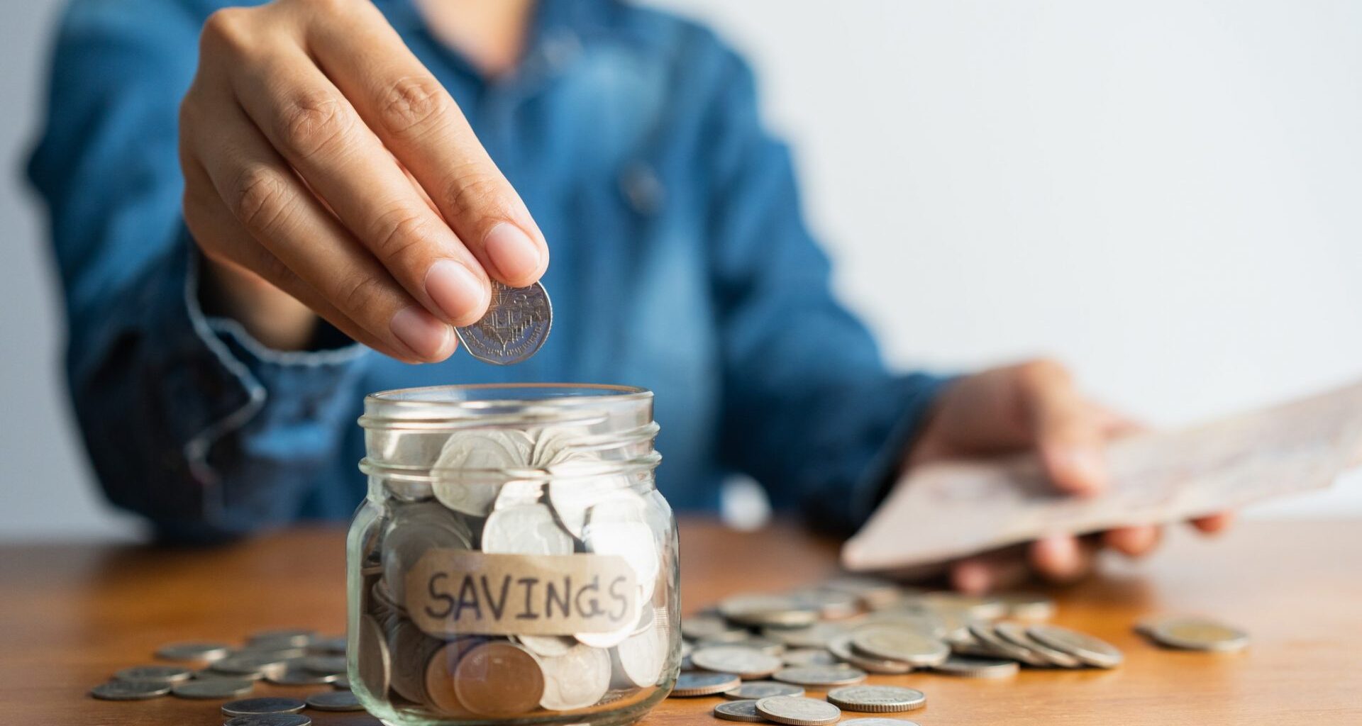 Woman hand is putting a coin into a glass bottle next to a pile of coins on a brown wooden table