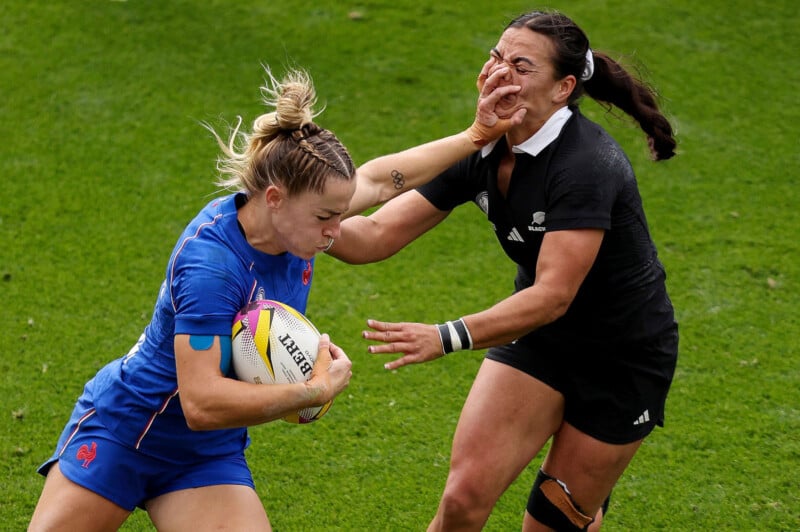 A rugby player in a blue uniform carries the ball while fending off an opponent in a black uniform with an outstretched arm. Both are focused and determined on a grassy field.