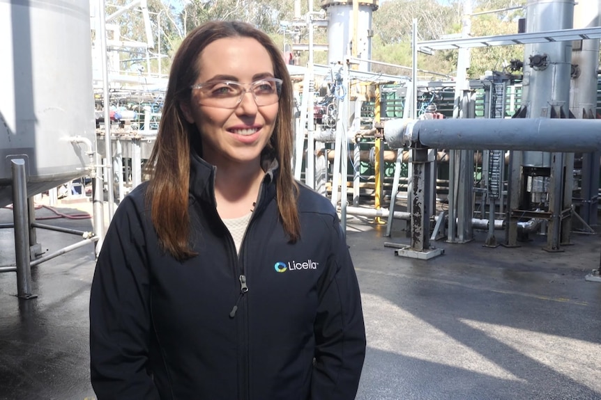 A woman wearing a navy blue jacket and safety goggles, she is standing in front of a tank and pipes.