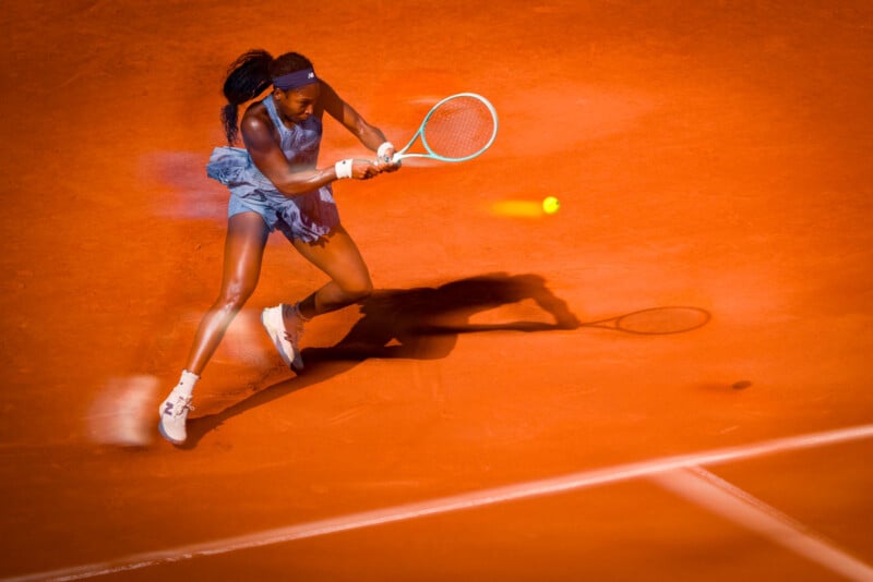A female tennis player in a blue outfit hits a backhand shot on a clay court, her shadow and the tennis ball visible on the orange surface.