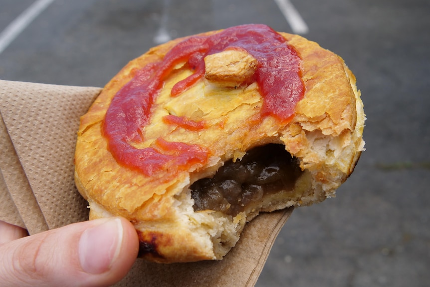 A bite has been taken out of a golden yellow pastry pie drizzled with tomatoe sauce, showing the meat inside.