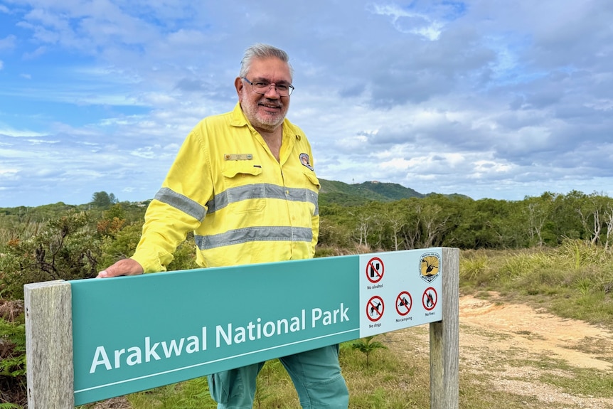 A man stands next to a national parks sign with the Cape Byron Lighthouse in the background