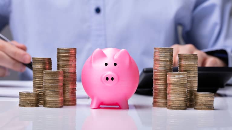 A little pink piggy bank between stacks of coins with a person seen in the background holding a pen and using a calculator.