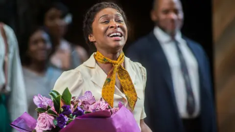 Getty Images Cynthia Erivo smiles at the end of a performance of The Color Purple on Broadway in 2014. She is wearing a cream blouse, yellow neckerchief and is holding a purple bouquet.