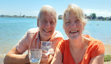 Chris and Ruth Stone-Houghton sit on a picnic bench, smiling into the camera. They are holding alcoholic drinks, and behind them is a body of water