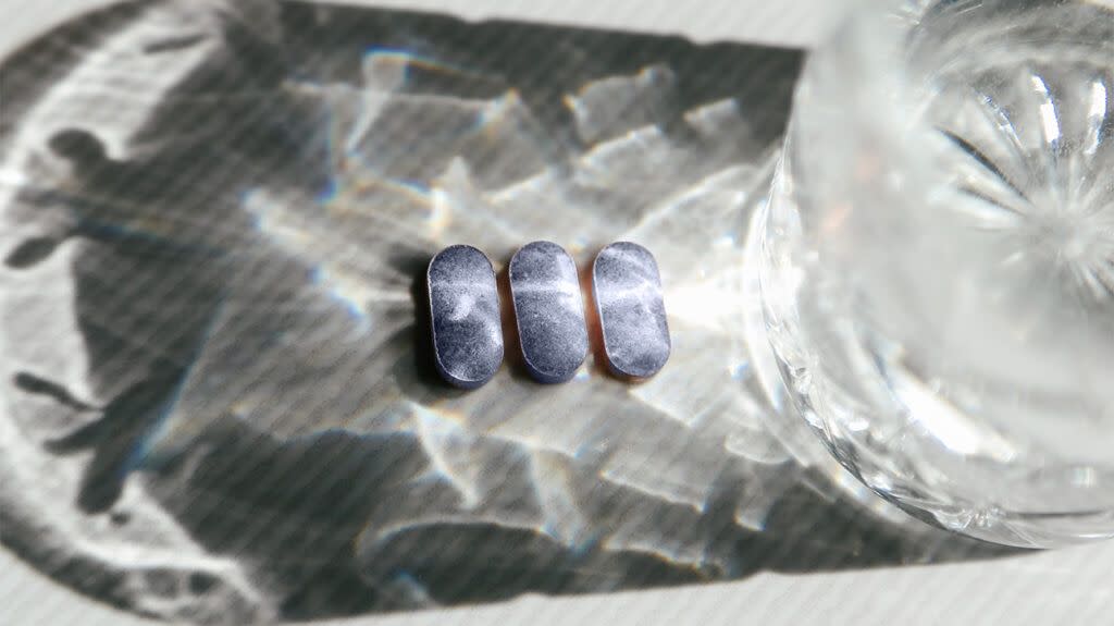 Three supplement pills next to a glass of water and its reflection