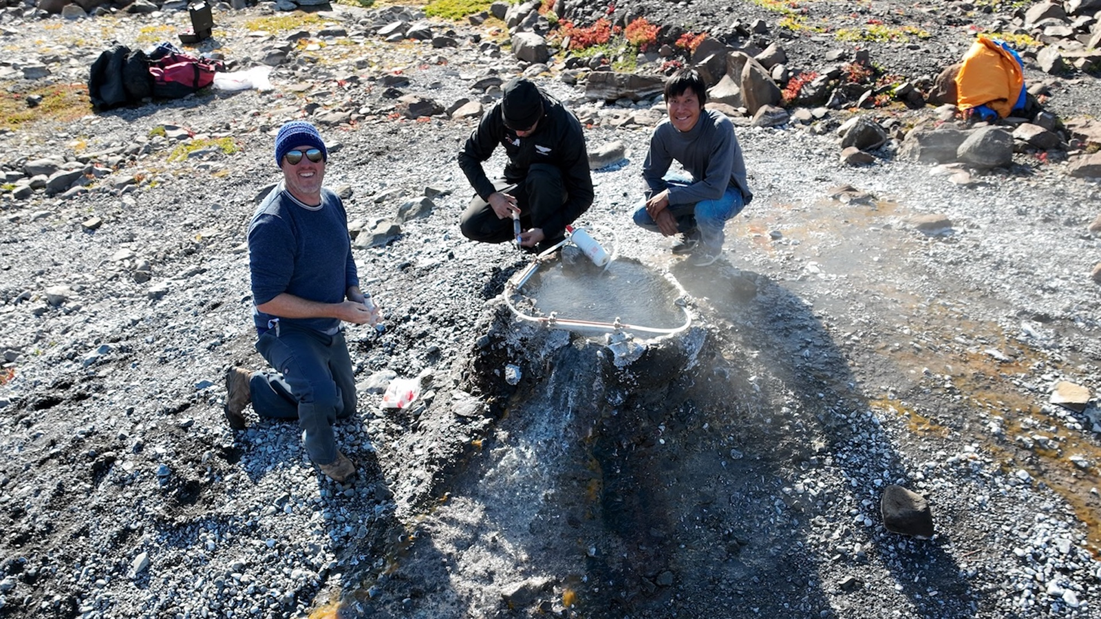 A photo of three men crouched around a hole in the ground emitting helium gas