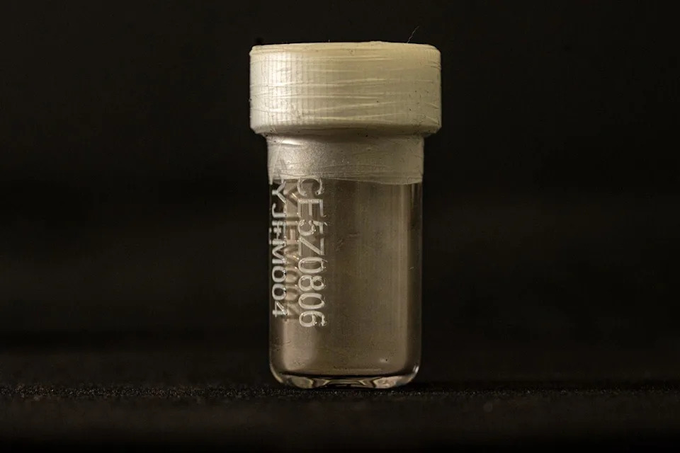 A small clear vial with a white plastic top and tape around the lid. It is lit against a black background. On the front is a code. Inside at the bottom there is a coating of dust.