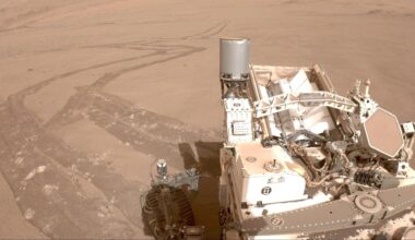 An image taken from one of Perseverance's cameras with the rover in the bottom right of the image and a long windy trail of tire tracks in the reddish brownish dirt of Mars behind it