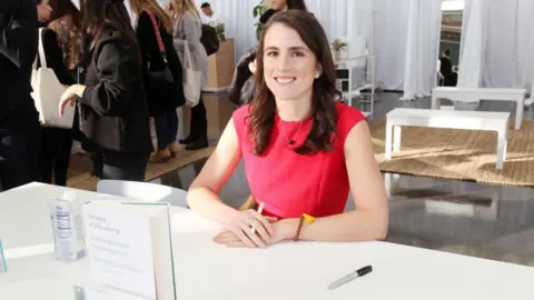 Getty Images Tatiana Schlossberg with long brown hair and wearing a red short-sleeved dress, sits behind table where her book  is stood up with hands crossed holding a pen
