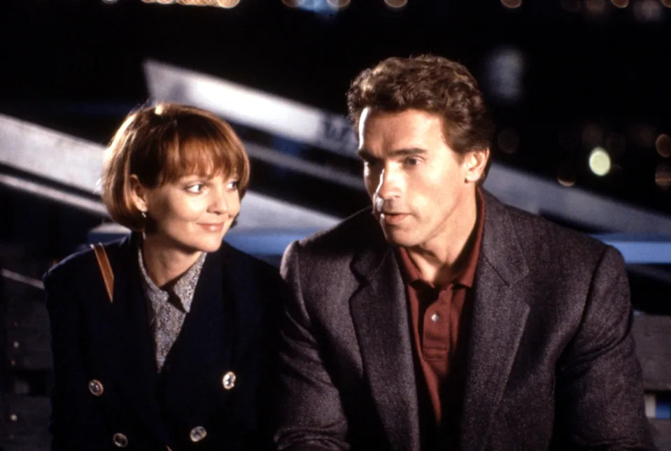 A man and woman sit together, the woman with a bob haircut smiling at the man. The man is wearing a blazer over a polo shirt, looking pensively ahead