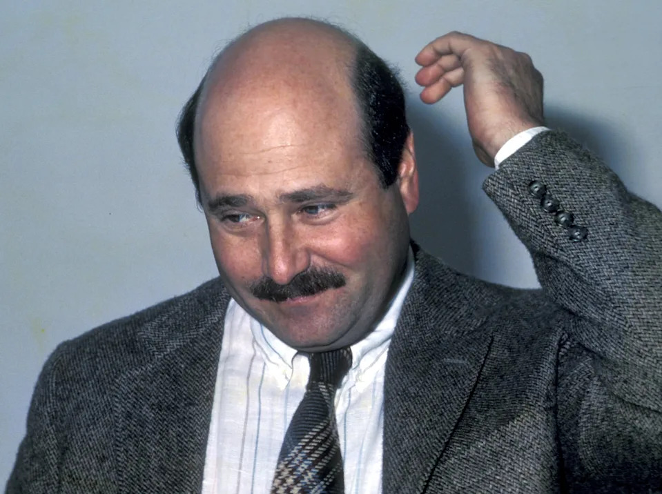 Bald man in a suit and tie with a mustache, scratching his head with a thoughtful expression