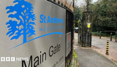 Main gate of St Andrew's Healthcare. There is a metal sign on black railings, with blue logo shaped like a tree. To the right is an iron fence and some black and yellow striped bollards.