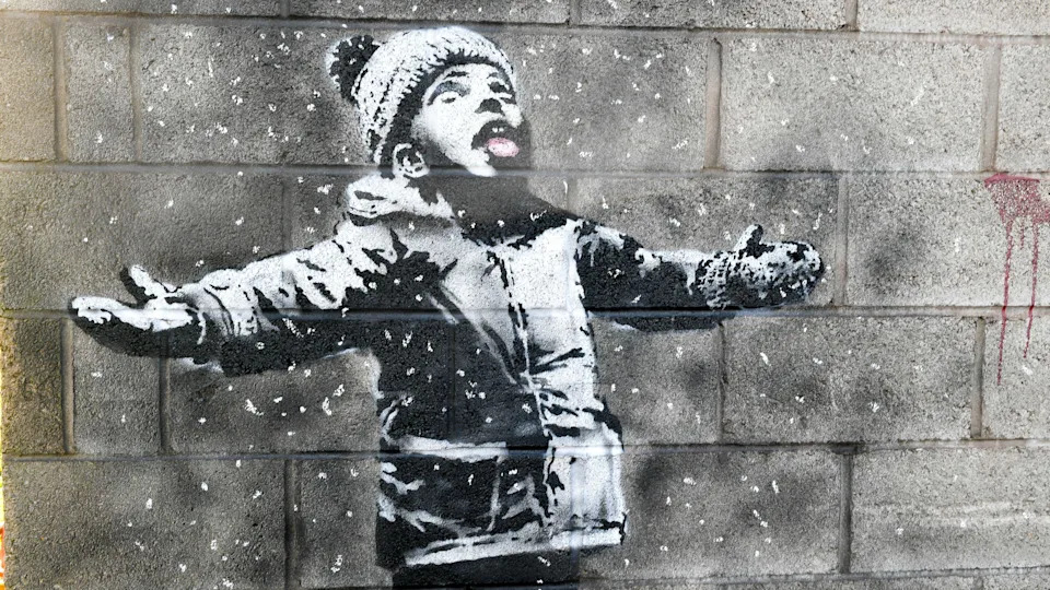 A lack and white mural of a small boy wearing a woolly hat, looking up with his mouth open, catching snowflakes