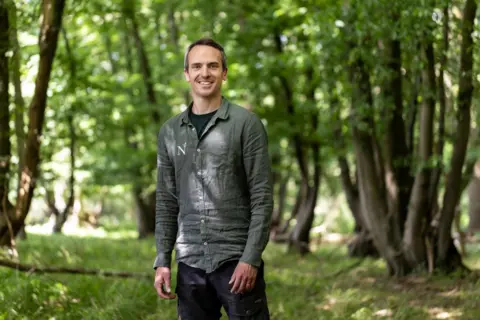 Jonathan Perugia for Nattergal A man stands in a green long sleeve shirt and black trousers, in the middle of some woodland. He is smiling at the camera.