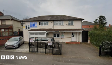 General view of the entrance of Naseby Medical Centre in Birmingham. A cream coloured building with a small driveway outside. Two cars are parked in the driveway which has some iron gates across part of it.