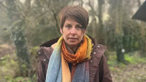 Lisa Morrison, a woman with short, brown hair, looks directly into the camera with a neutral expression.  She is standing in a wooded area, wearing a brown hooded jacket and multicoloured scarf. 