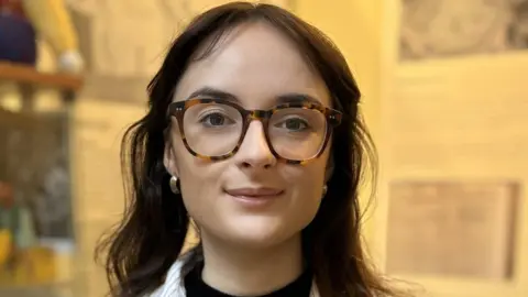 Head and shoulders image of Isla Kinear from the Fintry Development Trust. She has long brown hair and is wearing large tortoise-shell frame glasses and gold hoop earrings
